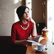 Женщина в красной кофте читает книгу у окна, наслаждаясь кофе.